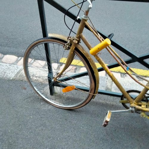 Photographie en couleur prise avec un téléphone portable. On y voit un vélo doré un peu vieillot attaché à une barrière sur un trottoir. Le pneu avant est complétement dégonflé. C'est nul.